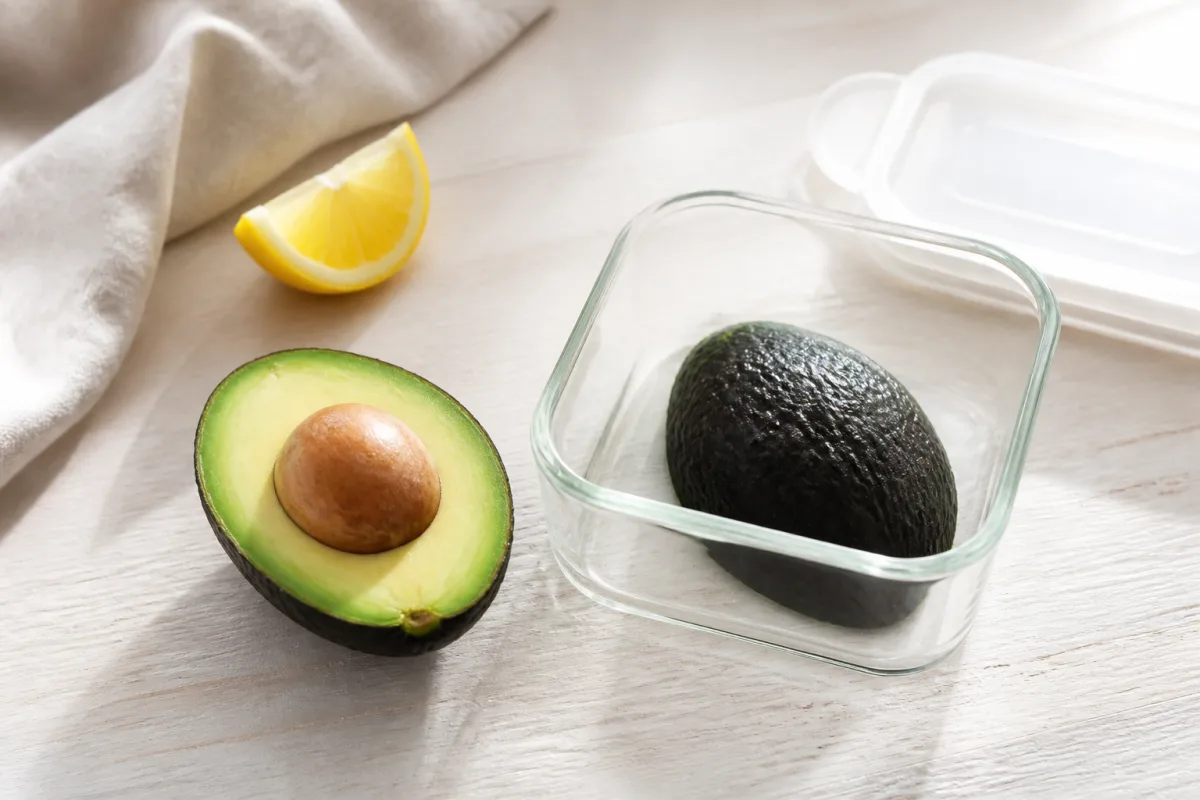 Fresh avocado half with its pit beside a lemon wedge and a glass storage container holding the remaining avocado half.